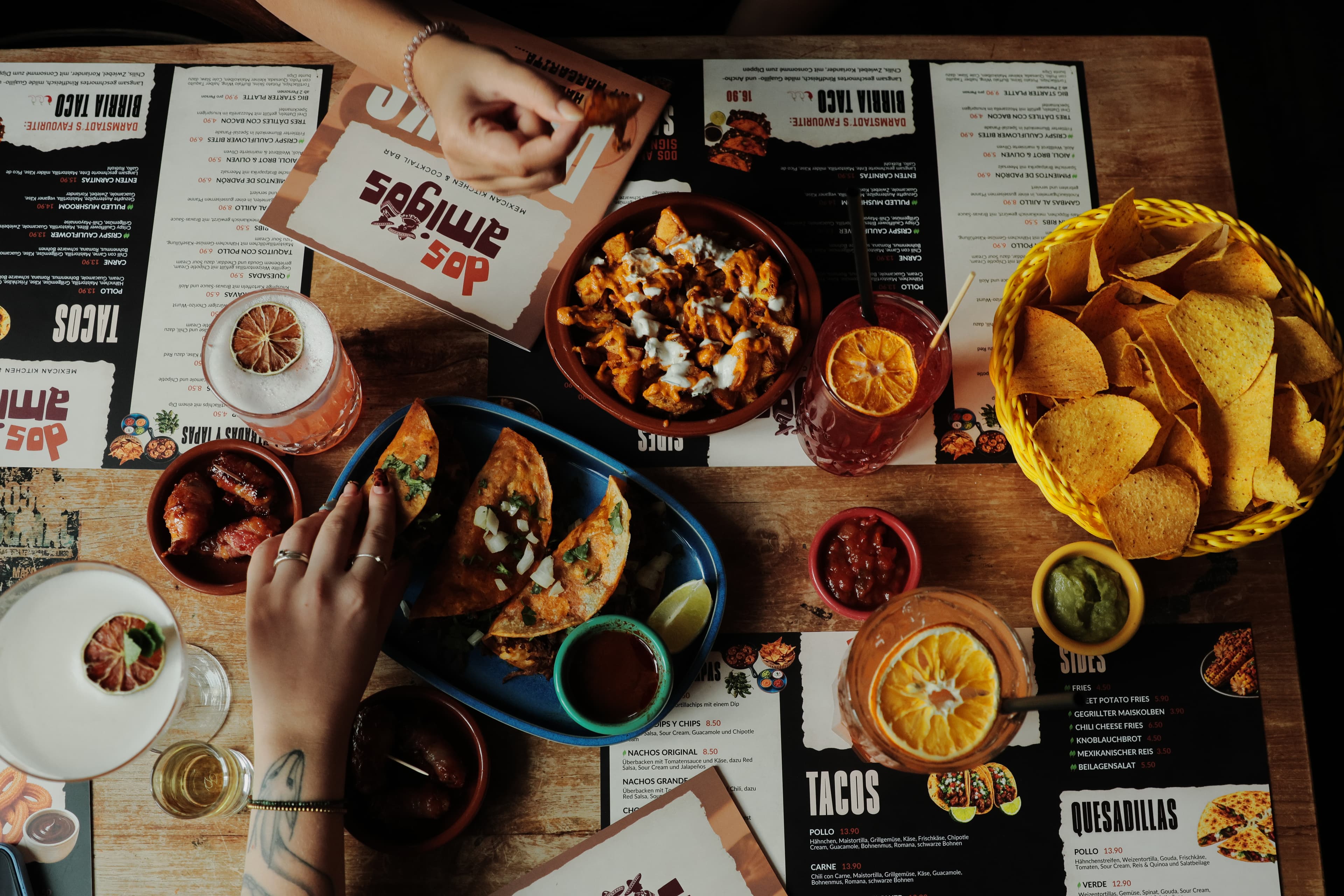 Eine Gruppe Freunde lacht gemeinsam an einem Restauranttisch bei Lichterketten, umgeben von Tacos, Margaritas und geteilten Speisen